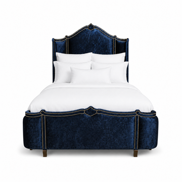 A luxury bed with a tall, curved headboard and matching footboard upholstered in deep navy blue crushed velvet, accented with antique gold nail head trim. All Haute House products are built in the United States and customizable 